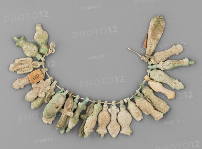 Fragment of a necklace of amulets 26 pieces, Goluchów Greater Poland Voivodeship; amulet necklace composed of 26 beige to greenish pieces including tit knots, ankh life signs and various objects; grave goods; ca. 1069–656 BC, Third Intermediate Period, Egypt; amulet, jewelry, A curved string holding a group of small pendant-like objects arranged like a garland, each pendant having a rounded, elongated, or slightly bulbous shape with small loops at their tops; the objects show a weathered, rough surface texture with areas of pitting and encrustation. The colors visible include shades of green, pale green, light tan, beige, off-white, and a few spots of darker brown; a small piece of twine or cord threads through the loops and appears off-white, and a short length of thicker twisted cord is tied near one end. The pendants vary slightly in shape and size and are spaced closely together along the cord, fanning outward to form a semicircular layout on a flat, smooth gray background.