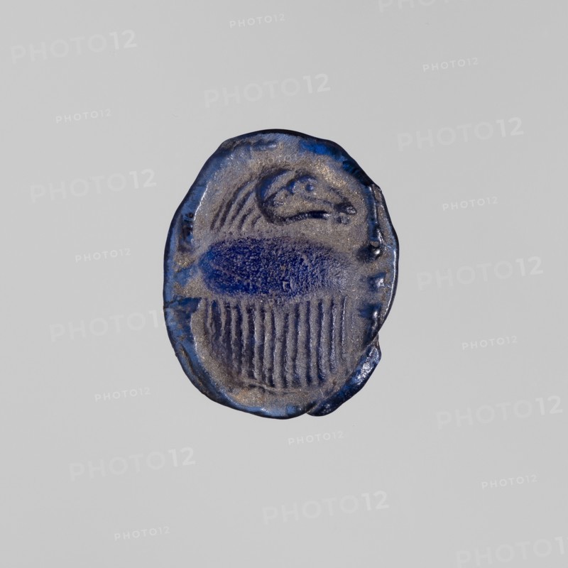 Scarab with a ram's head, ram's head ornament, sacred animal protective amulet in navy blue glass; scarab amulet, height 2.4 cm, width 1.9 cm; 664–525 BC, Late Period, 26th Dynasty, Egypt;, A small oval object with irregular edges photographed against a plain light gray background; the object has a textured surface in shades of deep blue and metallic gray, with a darker blue central oval area and vertical striations beneath it, and a rounded raised form toward the top that suggests a curved shape.