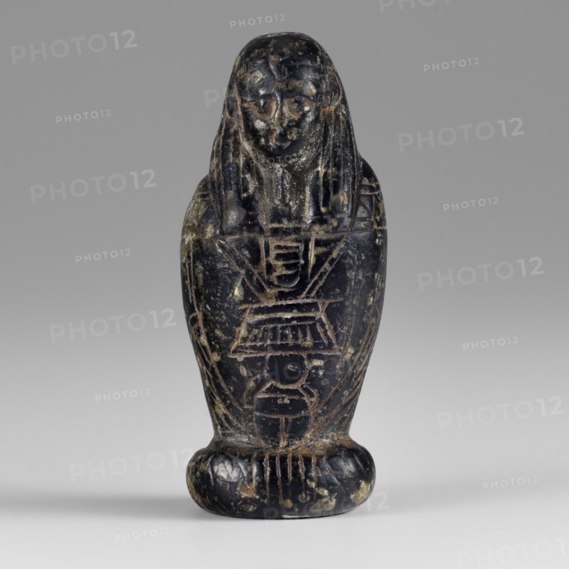 Amulet - god Osiris in hydria; maker, Osiris Canopic mythology; Egyptian deities; Egyptian religion; 1st–2nd century AD, Roman period, Egypt; amulet; height 4.9 cm, width 2.1 cm, A small carved figure standing upright on a rounded base, with a stylized human-like face and long hair or headdress that falls to the shoulders; the body tapers downward and is inscribed with linear carved motifs and geometric patterns. The surface shows worn areas and mottled spots, with visible scratches and chips. The dominant colors are dark black with lighter beige or tan wear marks and some gray tones in the background.