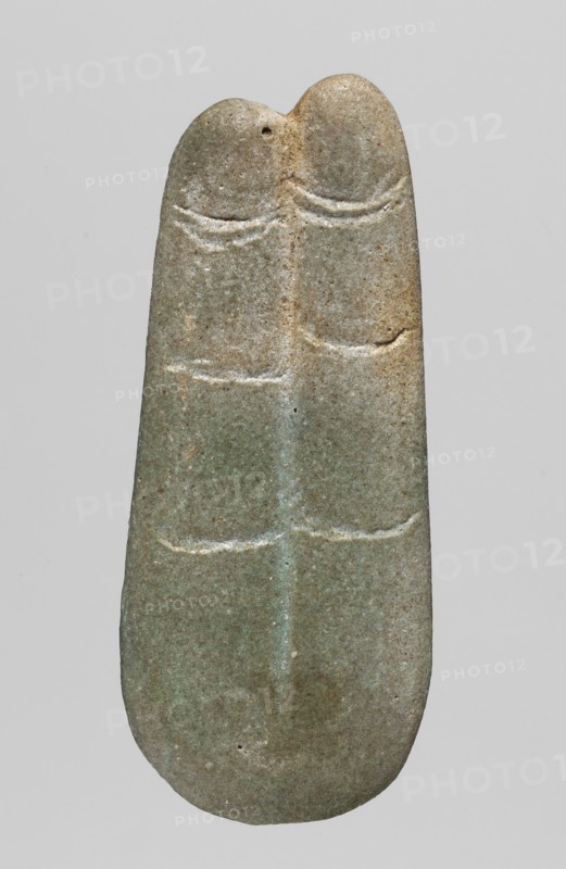 Amulet — two fingers; maker, Goluchów Greater Poland Voivodeship; fingers; amulets — body parts; grave equipment; 664–525 BC; Late Period, 26th Dynasty, Egypt; amulet; height 5.5 cm; width 2.4 cm, A single elongated, rounded object resembling a carved stone is centered against a plain light gray background, showing a muted palette of greenish-gray with areas of beige and brown. The stone has a smooth surface with subtle weathering and speckled texture, and a vertical ridge runs down its middle dividing the piece into two symmetrical lobes at the top. Shallow, curved incisions form parallel horizontal lines across the upper and middle sections on both sides of the central ridge, and a small circular hole is visible near the top left lobe. The overall shape tapers slightly toward the top and rounds more at the bottom, and the stone displays variations of color from pale green to sandy tan with faint lighter highlights along the carved grooves.