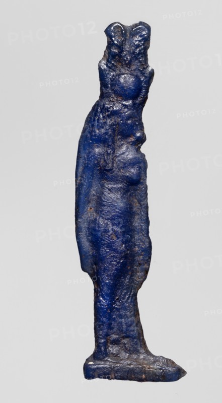 Amulet - goddess Hathor; Dark blue glass with a touch of violet; Ptolemaic Period, 332–30 BC, Egypt; amulet; height 5.4 cm, width 1.1 cm; depicts Hathor Egyptian goddess., A slender vertical object shaped like a stylized standing figure with an elongated body and a suggestion of shoulders and hips, rendered in a deep blue color with patches of darker and lighter blue and small areas of brownish discoloration, showing a rough, uneven surface texture with visible bumps and irregularities, standing on a narrow flat base that is also blue with worn edges, all set against a plain light gray background.