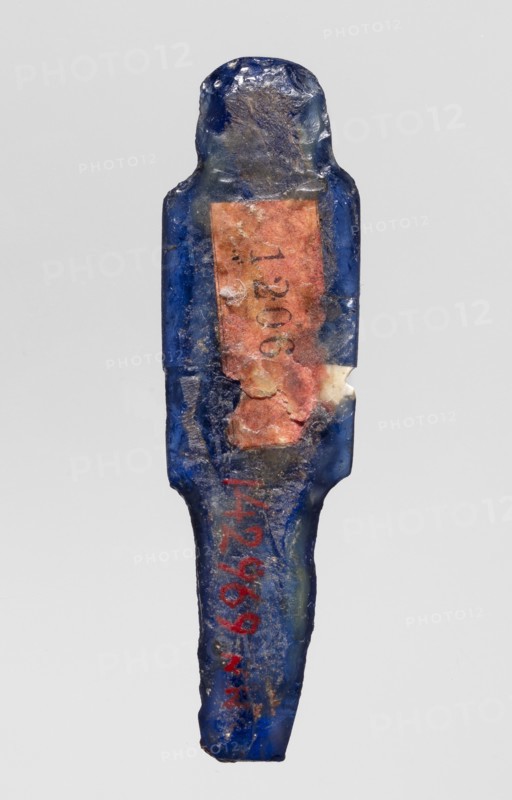 Amulet — god Horus; dark blue glass; Ptolemaic Period, 332–30 BC, Egypt; amulet; height 4.6 cm, width 1.3 cm; Egyptian deities; Egyptian religion., A single elongated object with an irregular, roughly rectangular top and a tapered bottom, shown against a plain light gray background; the object is predominantly deep blue with areas of darker and lighter blue, and it has a worn, uneven surface texture with visible chips and rough spots; there is a central patch of muted orange-pink material adhered to the surface, with portions flaking and showing underlying blue and pale off-white material; there are traces of a reddish pigment on the lower portion and small areas of dark grayish residue across the object.