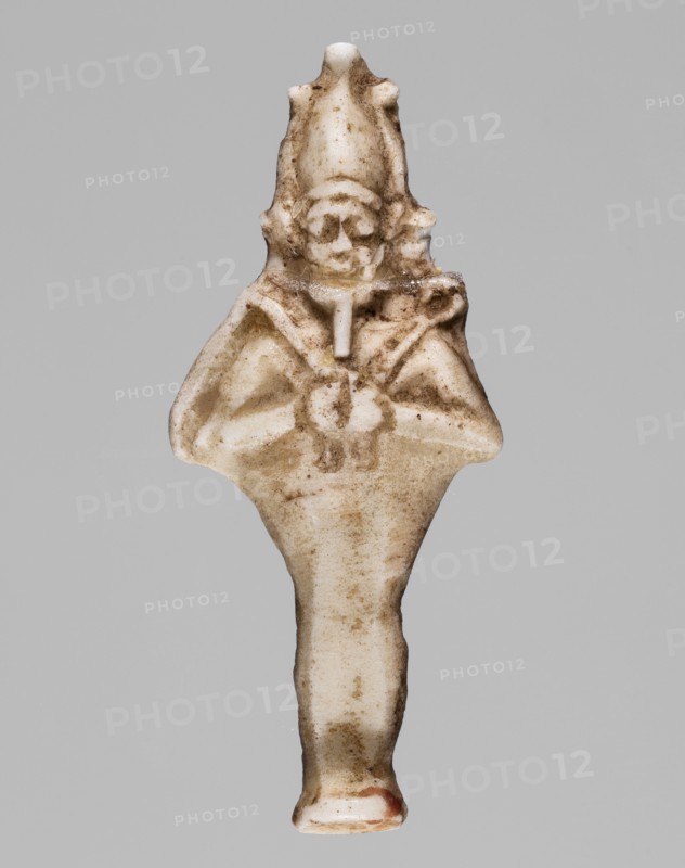Amulet — standing god Osiris; light yellow glass; Ptolemaic Period, 332–30 BC, Egypt; amulet; height 3.6 cm, width 1.6 cm; Egyptian deities; Osiris; Egyptian religion, A small carved figure in off-white and beige tones with brownish soil or age discoloration throughout, showing a stylized human shape with a pointed headdress, a rounded face area, and a suggested torso with arms folded or held together; the surface has rough and smooth areas with darker brown flecks and smudges, and a single small area of reddish-brown near the bottom; the background is a flat light gray.