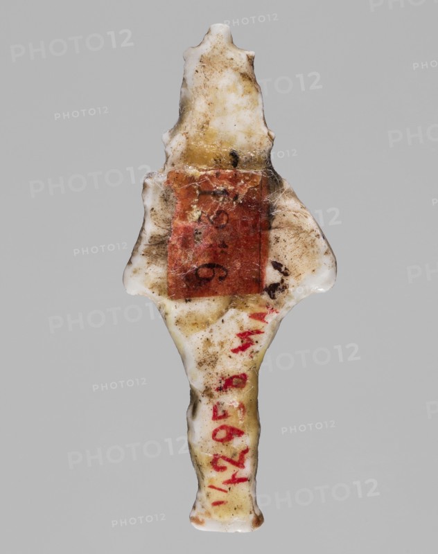 Amulet - standing god Osiris; Light yellow glass; 332–30 BC, Ptolemaic Period, Egypt; amulet; height 3.6 cm, width 1.6 cm; Egyptian deities; Osiris; Egyptian religion, An elongated, irregular object with a central narrow shaft and flared, wing-like projections near the top, appearing to be made of a hard, glossy material with a worn and dirty surface; the object shows a mix of off-white and yellowed areas with brownish grime and smudges, and a rectangular patch of muted reddish-brown material adhered near the upper middle, with traces of darker brown spots and fine fibrous debris around it; there are also several small red marks along the lower portion of the shaft, and the background is a uniform light gray.