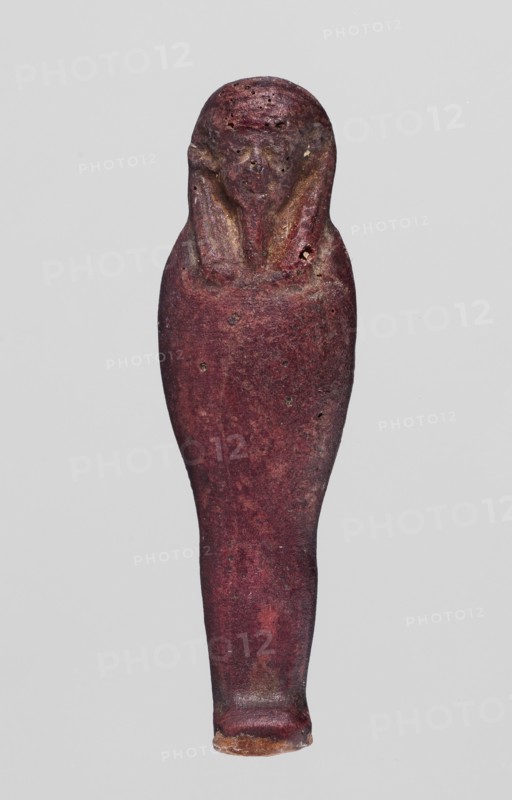 Amulet - mummy-shaped figurine;, Mummy-shaped figurine, back smooth; dark red glass; Ptolemaic Period, 332–30 BC, Egypt. Amulet; height 3.8 cm, width 1 cm., A single elongated figurine shaped like a wrapped human form with a rounded head and tapered body, shown against a plain light gray background. The surface of the object is predominantly deep reddish-purple with areas of darker maroon and some brownish patches. The head area shows subtle smoother contours and slightly lighter reddish-brown highlights, while the body displays a textured, worn surface with small pitted marks and faint darker spots. The lower tip of the object has a small exposed edge that is more brown and tan in color. Overall the object appears solid and slightly irregular in shape with a mix of reddish-purple, maroon, brown, tan, and faint gray tones.