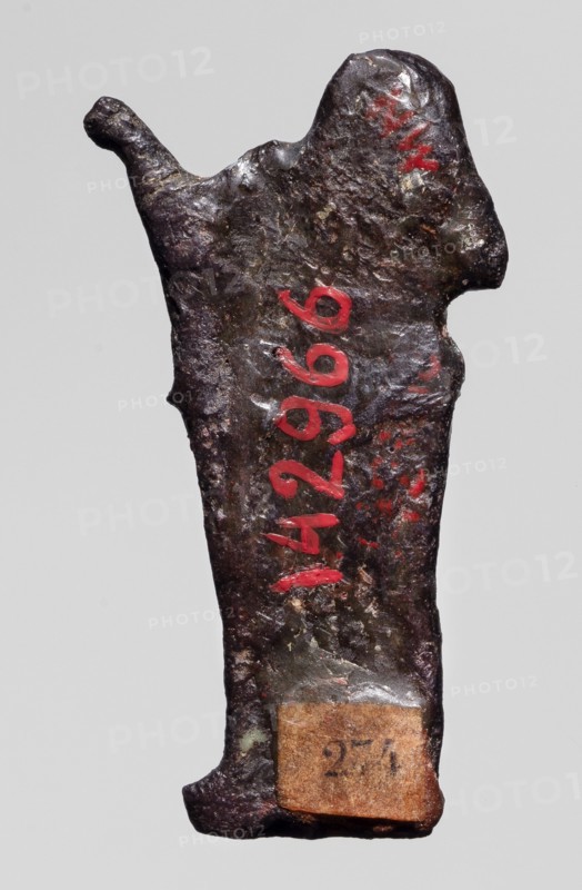 Amulet — goddess Seshat;, Dark brown glass; Ptolemaic Period, 332–30 BC, Egypt. Depicts Seshat Egyptian goddess associated with writing, harp iconography. Amulet; height 3.4 cm, width 1.7 cm., A roughly rectangular, irregularly shaped metal object with a rough, pitted surface is shown against a plain light gray background; the object has a dark brown to nearly black patina with areas of metallic sheen and hints of greenish corrosion, and there are a few strokes of bright red paint on its surface; the top edge is uneven with a protruding curved nub on one side and a rounded raised area on the other, the lower edge flares slightly and has a small attached rectangular fragment of brownish material with a worn appearance.