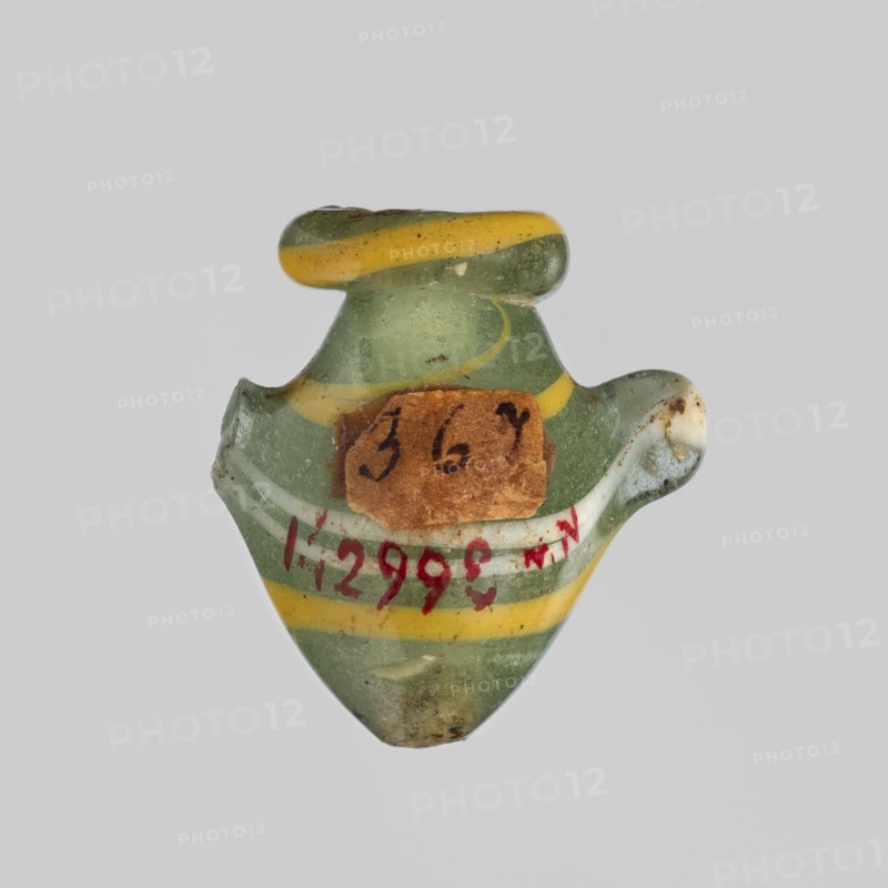 Amulet - heart ib, amulet, hearts ib representing a heart, amulet of a body part, grave equipment, New Kingdom, 18th Dynasty, ca. 1550–1295 BC, Egypt; height 2.2 cm, width 1.6 cm, A small glass object with a translucent green body and a few curved yellow bands, shaped like a tiny vessel with a rounded top and a wider flared midsection, showing some chips and rough edges and a small brown paper fragment stuck on one side; the surface has a few darker spots of dirt and wear and the colors visible are green, yellow, brown, white, and hints of red.