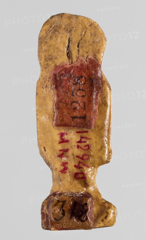 Amulet - tit, tit knots, protective amulet, grave good, with hieroglyphs, yellow glass; Ptolemaic Period, 332–30 BC, Egypt; amulet, height 3.3 cm, width 1.2 cm., A small carved object resembling a stylized human figure viewed from the back, with a smooth, worn surface in a yellow-beige color mottled with brown spots and darker speckles, a reddened rectangular patch near the upper back area, a few strokes of red pigment along the spine area, and a darker brown rounded base area; the background is a neutral light gray.