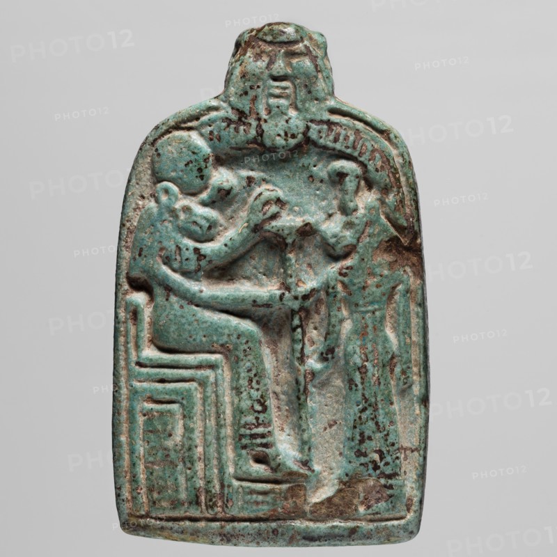 Amulet — stele with lion-headed goddesses Bastet and Sekhmet?, lionesses, Egyptian deities; possibly Sekhmet, Bastet, Bes; thrones; ca. 747–525 BC 25th–26th Dynasty, Egypt; amulet; height 7.1 cm, width 4.6 cm., A carved rectangular amulet with rounded top showing a raised relief of a seated human figure facing a standing smaller figure, both rendered in shallow relief; the seated figure sits on a blocky, stepped seat while the standing figure reaches toward the seated figure; above them a larger head or mask-like form overlooks the scene; the surface shows a weathered, speckled patina in shades of turquoise and green with areas of darker brown and black spotting and some lighter worn patches; the background around the figures is slightly recessed and the edges of the object are smoothed and irregular.