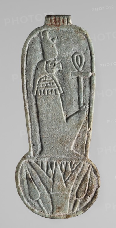 Amulet – weight of a menit necklace, An amulet in the shape of the counterweight of a menit necklace, with the image of a mummy-shaped, falcon-headed deity in a double crown holding an ankh in the longitudinal part and a lotus flower with two buds in the round lower part. On the reverse is a column of text with a sacrificial formula to the goddess Uadjit. Materials/icons: Re mythology, Uadjit, double crown, ankh, lotuses, hieroglyphs, sacrificial formulas, Egyptian deities. Date: 664–332 BC, Late Period, Egypt. Type: amulet. Dimensions: height 9.2 cm, width 3.3 cm., A vertical carved object with a rounded lower end, showing a raised profile of a human figure wearing a tall headdress and a patterned collar, holding a staff-like item; the surface is weathered and pitted with textured carving lines. The piece displays a palette of pale greenish-blue with areas of gray and small patches of brownish rust and darker green, with subtle variations across the relief and edges. The lower rounded...