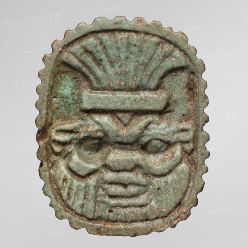 Amulet plaque with the head of the god Bes; plaque amulet, Egyptian deity Bes, wearing crowns. Late Period, Egypt, 664–332 BC. Height 3.4 cm; width 2.7 cm., A small oval object with a scalloped edge featuring a raised carved face with stylized facial features, including pronounced eyebrows, deep-set eyes, a broad nose, and a layered mouth area; the surface shows uneven texture with small pits and wear. The object displays a range of green tones from pale green to turquoise, with areas of brownish and reddish discoloration and darker green spots. The carving includes symmetrical vertical and curved ridges above the face that resemble a fan or headdress, and the relief varies in depth across the piece. The background behind the object is a smooth light gray.