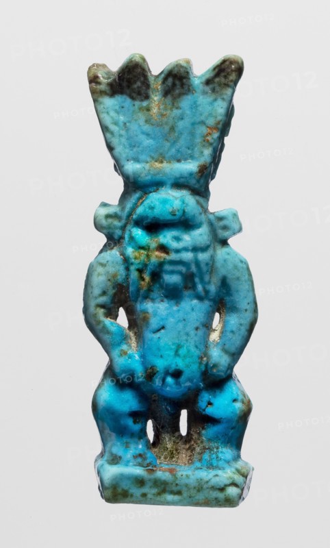 Amulet - god Bes, figurine of Bes standing on slightly crooked legs, with his hands resting on his thighs, wearing a crown of four feathers. Blue faience, feather tips black. 664–332 BC; Late Period, Egypt. Amulet; height 4.8 cm, width 1.8 cm., A small sculpted figurine colored predominantly bright turquoise with areas of darker teal and mottled brown-green patina, showing a stylized standing figure with a flared, crown-like headpiece and rounded shoulders, short arms bent at the sides, a rounded torso with subtle raised details, legs set apart above a rectangular base, and visible surface texture that includes small pits, worn edges, and irregular discoloration.