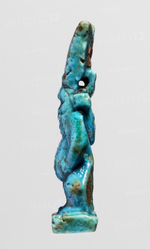 Amulet - god Bes;, Figurine of Bes standing on slightly crooked legs, hands resting on his thighs, wearing a crown of four feathers. Blue faience with black feather tips. Egyptian deities; feathers. Late Period, Egypt, 664–332 BC. Amulet; height 4.8 cm, width 1.8 cm., A small vertical sculpture shown against a light gray background resembling a stylized standing figure with simplified limbs and an elongated head, rendered in bright turquoise and aqua tones with patches of darker teal and green, and areas of brownish and rusty discoloration; the surface appears glossy and slightly irregular with a visible round hole near the upper portion, and the base is a small rectangular platform in similar turquoise shades.