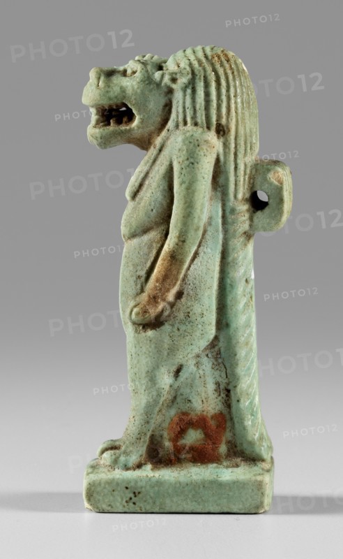 Amulet - goddess Toeris, Toeris myth., Egyptian deities, crocodiles, hippopotamuses. Figurine of the goddess Toeris in the form of a pregnant hippopotamus standing on lion legs and equipped with a crocodile tail. A stand under the legs, a pendant on the back. 664–332 BC; Late Period, Egypt. Amulet; height 5 cm, width 1.5 cm., A small statuette of a standing anthropomorphic lion figure rendered in a pale green-blue glazed material, showing a mane with vertical grooves, a snarling open mouth, and a defined nose and eyes; the figure’s arms hang down along its sides and it stands on a rectangular base. The surface shows areas of darker green and brown discoloration and a patch of reddish-brown staining near the lower front. The background is a smooth mid-gray that transitions to a lighter gray toward the bottom.
