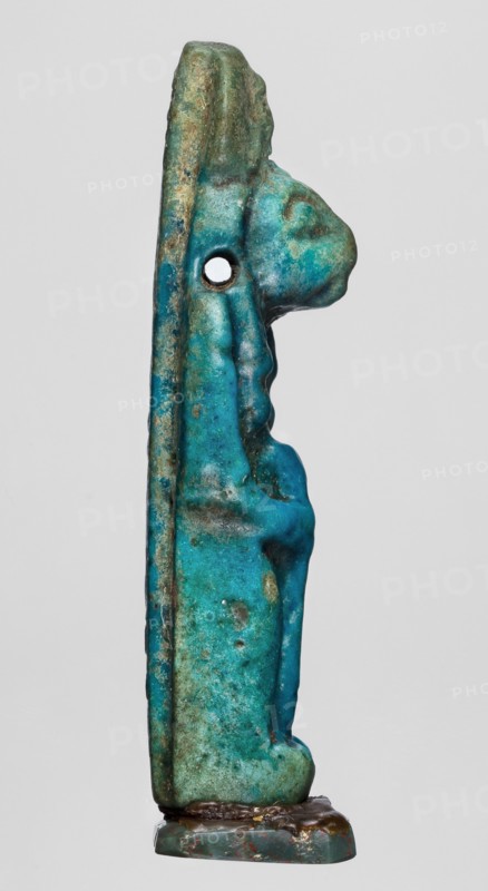 Amulet — figurine of a lion-headed goddess Sekhmet?, possibly Wajit; Egyptian deity. Sekhmet with a solar disk with a uraeus on her head, standing with a lotus scepter in her right hand left lowered. Late Period, 664–332 BC, Egypt. Amulet; height 5.5 cm, width 1.8 cm., A vertical, narrow carved object shown in profile with a patinated surface in shades of turquoise, teal, green, and hints of brown and blue, featuring a rounded protrusion near the top and an oval hole through the mid-upper area, mounted on a small rectangular base with darker brown and green tones; the surface shows uneven texture, weathered spots, and subtle raised contours suggesting carved details.