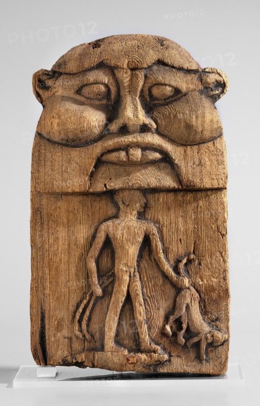 Magical stele topped with the mask of the god Bes. Magical stele cippus depicting in the lower register a naked Horus-Child in a scene of triumph over dangerous creatures. In his right hand, Horus likely holds two snakes, in his left a four-legged animal—perhaps a hyena or a lion. Horus stands on a crocodile. The depiction is related to the myth of a venomous snake attacking the infant Horus during the absence of his mother, Isis; the initiator of the attack was Seth, brother of Osiris, whom young Horus fought to avenge his father's death. The upper part of the monument depicts the face of the god Bes. 664–30 BC; Late Period–Ptolemaic Period, Egypt. Stele; height 20.5 cm, width 12.5 cm., A carved wooden plaque showing a large stylized face at the top with pronounced cheekbones, a broad nose, full lips, and deep-set eye shapes, rendered with visible carving marks and darkened recessed lines; below the face a carved human figure stands with an elongated torso and limbs, one arm extended ...