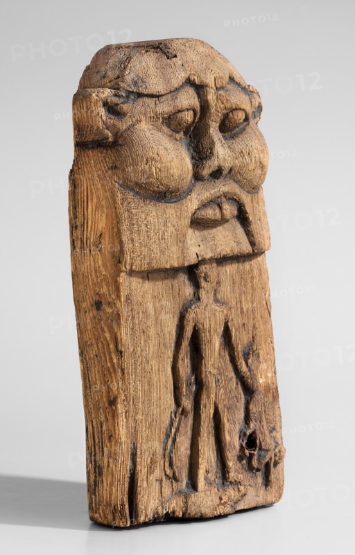 Magical stele topped with the mask of the god Bes. Magical stele cippus depicting in the lower register a naked Horus-Child in a scene of triumph over dangerous creatures. In his right hand, Horus likely holds two snakes; in his left a four-legged animal—perhaps a hyena or a lion. Horus stands on a crocodile. The depiction relates to the myth of a venomous snake attacking the infant Horus during the absence of his mother Isis; the initiator of the attack was Seth, brother of Osiris, whom young Horus fought to avenge his father's death. The upper part of the monument depicts the face of the god Bes. Date: 664–30 BC Late Period–Ptolemaic Period. Material/object type: stele. Dimensions: height 20.5 cm; width 12.5 cm., A carved wooden figure shown upright against a pale gray background, with visible grain and textured tool marks; the top portion features a stylized face with pronounced rounded cheeks, deep-set eye hollows, a broad nose, and a downturned mouth, all carved in relief and show...