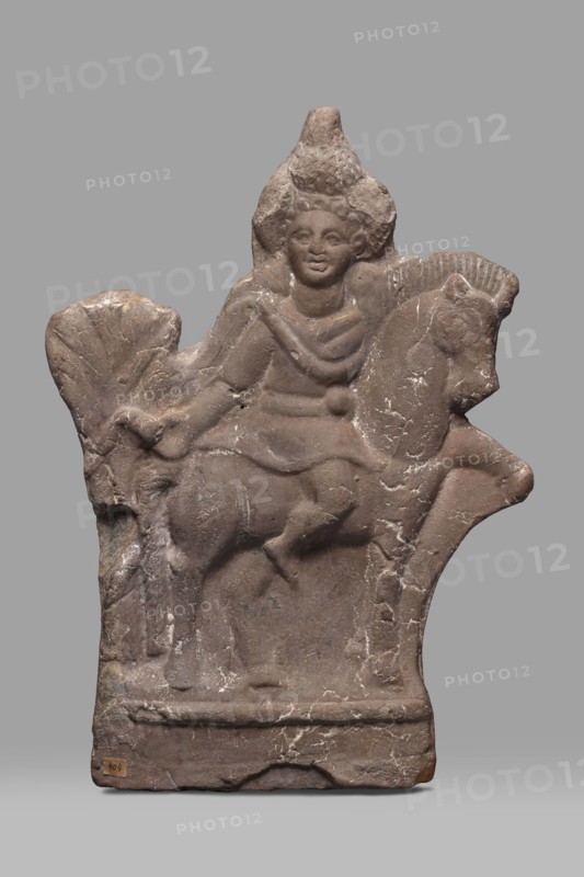 Figurine of Harpocrates-Heron on horseback, figurine of a deity sitting on a horse with a wreath and a double crown on his head, dressed in a short military tunic and cloak. The horse is walking to the right, the deity is facing the viewer, to the right. Behind the horse's rump is a palm tree, 2nd century, Egypt, height 29 cm, width 19.3 cm, Egyptian deities, Harpocrates mythology, Heron mythology, horses., A weathered stone relief in shades of brown and gray shows a carved figure seated on an animal resembling a horse. The figure has a rounded face and stylized hair or headdress and wears draped garments suggested by carved folds across the torso. The animal has a defined head with a visible ear and snout and a molded mane. The relief surface displays areas of lighter discoloration and small cracks, with rough, chipped edges along the top and sides. The base is rectangular and slightly raised, with worn contours and patches of lighter mineral streaking. The background behind the carvi...