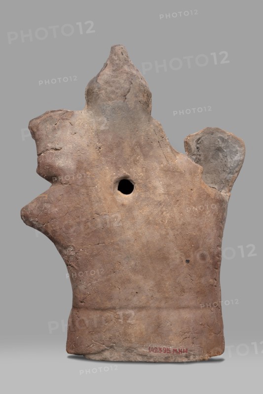 Figurine of Harpocrates-Heron on horseback, Egyptian deity Harpocrates combined with Heron, deity seated on a horse wearing a wreath and a double crown, dressed in a short military tunic and cloak. The horse walks to the right while the deity faces the viewer to the right; a palm tree appears behind the horse's rump. 2nd century, Egypt; terracotta figurine, height 29 cm, width 19.3 cm., A weathered clay object with an irregular, roughly vertical silhouette and an overall earthy brown color with patches of gray and lighter beige, showing a rough, pitted surface texture and uneven edges; a rounded protrusion on one side and another irregular lobed extension near the top, plus a central circular hole; the base is broader and slightly darker with subtle surface cracks and a few small darker spots; the background is a flat gray.