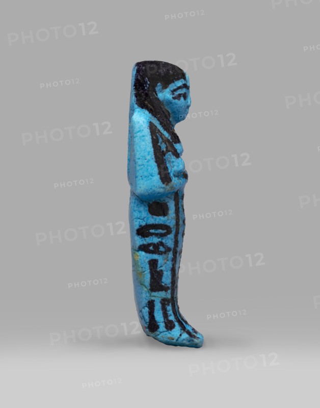 Queen Henuttawi's shabti, Henuttawi queen of Egypt; ca. 1100–1000 BC. Mummy-shaped funerary figurine made of blue faience with dark-painted details. Hands crossed on the chest, right folded over left; each hand holds a hoe. Three-part smooth wig with border and uraeus above the forehead. Round face with clearly marked eyes and eyebrows. Wide bracelets on the wrists and a necklace with a silver ornament on the chest. Facial features, wig, tools and jewelry painted with black dye. Painted inscription in three columns on the sides and in front of the legs reading: Ushabti of Osiris, the king's wife, Henut-tawi, with Osiris, the great god. ca. 1069–ca. 945 BC; Third Intermediate Period, 21st Dynasty; Deir el-Bahari . Height 12 cm; width 4.6 cm., A small vertical figurine in bright turquoise blue with areas of dark black paint, shown in profile against a neutral gray background; the figure has a rounded head with black-painted hair and facial markings, a slender body with the arms held clos...