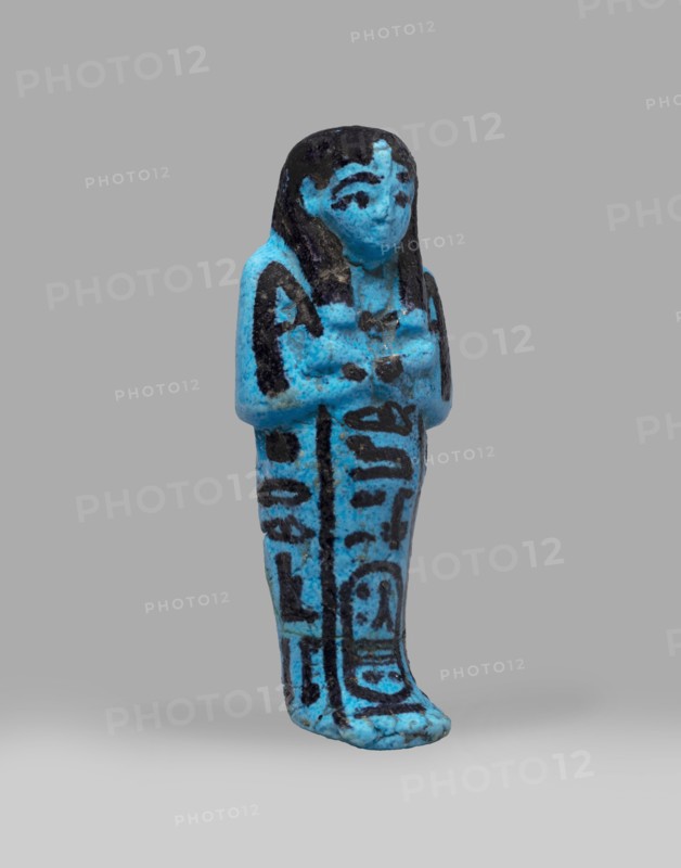 Queen Henuttawi's shabti, Henuttawi queen of Egypt; ca. 1100–1000 BC. Mummy-shaped funerary figurine made of blue faience with details in a dark color. Hands crossed on the chest, right folded over the left; both hands hold hoes. Wears a smooth three-part wig finished with a border and a uraeus above the forehead. Round face with clearly marked eyes and eyebrows. Wide bracelets on the wrists and a necklace with a silver ornament on the chest. Facial features, wig, tools, and jewelry painted with black dye. Painted inscription arranged in three columns on the sides and in front of the legs reading: Ushabti of Osiris, the king's wife, Henut-tawi, with Osiris, the great god. Attributed ca. 1069–ca. 945 BC; Third Intermediate Period, 21st Dynasty; shabti. Height 12 cm, width 4.6 cm., A small standing figurine rendered in bright blue with black painted details, showing a stylized human form with long hair, a defined face, and arms crossed over the chest; the surface has a slightly rough, ag...