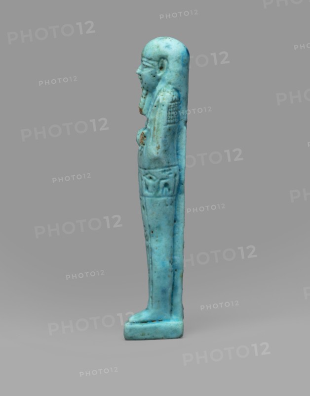 Ushabti Wahibre-em-achet, Back pillar, hieroglyphs, Wahibre-em-achet ca. 664–525 BC. Mummy-shaped funerary figurine shabti. Made of blue faience with black details. Stands on a base, supported by a pillar at the back. Arms crossed on the chest, right folded over left. From under the shroud only the hands clenched into fists are visible. Right hand holds a hoe, left a pickaxe; a basket is slung over the left shoulder. Engraved tools. Smooth three-part wig with front strands longer than the back. Straight, braided beard. Large eyes and a prominent mouth with a slight smile. Engraved inscription arranged in a T-shape: one line running around the figure at hip level and one column on the front of the legs reading, Shd Osiris the father of God, priest ... Uah-ib-Re-em-achet born of Ta-net-Amun. Date: 664–525 BC, Late Period, 26th Dynasty, Egypt. Dimensions: height 13.4 cm, width 3 cm., A small upright figurine depicted in profile, rendered in shades of blue and turquoise with some darker bl...