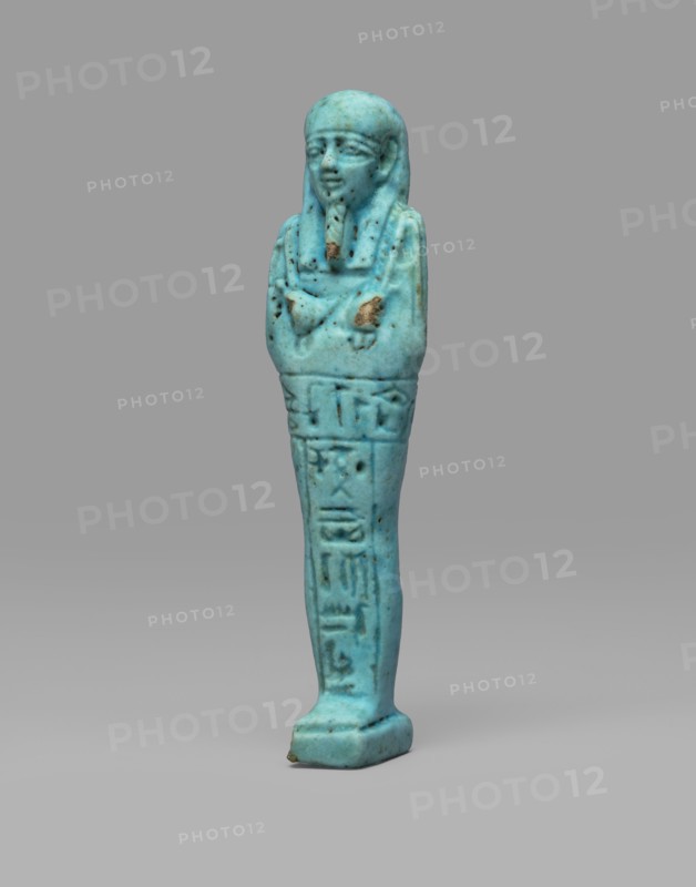 Ushabti Wahibre-em-achet. Mummy-shaped funerary figurine shabti, ca. 664–525 BC, Late Period, 26th Dynasty, Egypt. Made of blue faience with black details. Stands on a base, supported by a pillar at the back. Arms crossed on the chest, right folded over left. From under the shroud only the hands clenched into fists are visible; right hand holds a hoe, left a pickaxe; a basket is slung over the left shoulder. Engraved tools. Smooth three-part wig with longer front strands than the back. Straight, braided beard. Large eyes and a mouth stretched in a slight smile. Engraved inscription arranged in a T-shape — one line running around the figure at hip level and one column on the front of the legs: Shd Osiris the father of God, priest ... Uah-ib-Re-em-achet born of Ta-net-Amun. Height 13.4 cm; width 3 cm., A small turquoise-blue figurine shaped like a standing mummiform person with a rounded head and stylized facial features, crossed arms, and a long narrow body; the surface shows some darke...