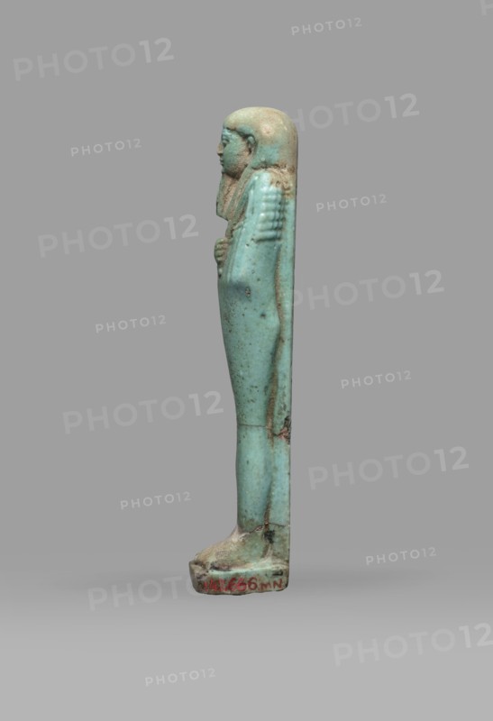 Ushabti Psamtyk-seneb, Wigs, funerary figurine, hieroglyphs, beards facial hair, Psamtyk-seneb ca. 747–332 BC. Mummy-shaped figurine, standing on a base, back supported by a pillar/pilaster. Arms crossed on the chest, right folded over left. Right hand holds a long-bladed hoe, left a pickaxe; handles of both hoes are rolled up at the ends. A bag is slung over the left shoulder on a single string. Wears a three-part smooth wig. Long tapering beard braided into a plait. Engraved inscription in one column on a pilaster: Osiris Psammetik-seneb born of Jerou justified by the voice. Date: 664–332 BC, Late Period, Egypt. Type: shabti. Height: 12.5 cm; width: 3 cm., A small upright figurine viewed in profile standing on a rectangular base, made of a smooth turquoise-blue material with areas of pale beige and brown discoloration; the figure has a rounded head with a close-fitting headdress that extends along the back, a defined facial profile, and a folded arm held against the chest; the body i...