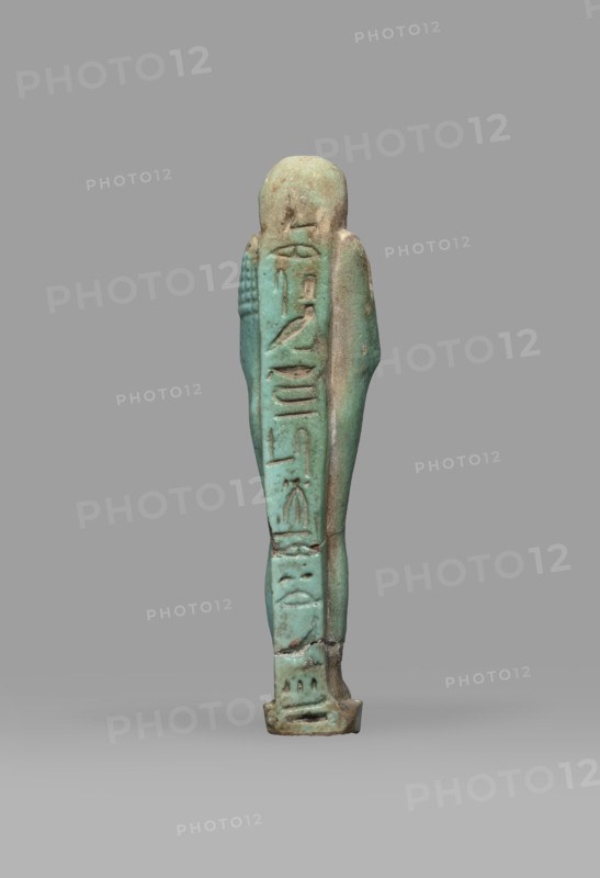 Ushabti Psamtyk-seneb, wigs, funerary figurine, hieroglyphs, beards facial hair, Psamtyk-seneb ca. 747–332 BC, back pillar/pilaster, grave goods, mummy-shaped figurine standing on a base, back supported by a pillar. Arms crossed on the chest, right folded over left. In the right hand a long-bladed hoe, in the left a pickaxe; handles of both hoes rolled up at the ends. A bag slung over the left shoulder on a single string on the back. Three-part smooth wig; long tapering beard braided into a plait. Engraved inscription in one column on a pilaster reading Osiris Psammetik-seneb born of Jerou justified by the voice. 664–332 BC; Late Period, Egypt. Shabti; height 12.5 cm, width 3 cm., A small upright figurine viewed from the back, with a slender elongated body and a rounded head, standing on a short rectangular base; the surface shows a weathered, slightly rough texture with visible chips and small cracks; the left side of the figure has a series of incised vertical shapes and shallow reli...
