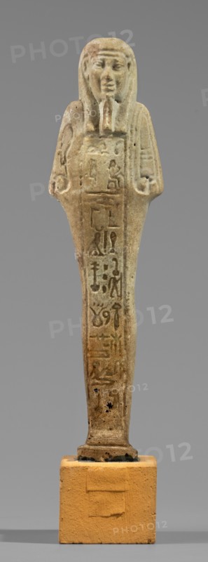 Hornakht's shabti, back pillars, hieroglyphs, wigs, beards facial hair. Hornakht ca. 525–30 BC. Funerary figurine mummy-shaped shabti, standing on a base, back supported by a pillar/pilaster. Arms crossed on the chest, right folded over the left. In the right hand a long-bladed hoe and two strings from which hangs a semicircular basket on the back, slightly above the elbow; in the left hand a whip. Hands and tools engraved. Long, three-part smooth wig; long straight beard of uniform width. Face with coarse features, roughly defined eyes, wide nose, prominent mouth set in a slight smile. Right ear significantly larger than the left. Inscription carved in a single column on the front from chin to feet, naming the deceased Hornakht and his mother content otherwise unclear. Date: 664–30 BC Late Period–Ptolemaic. Egypt. Shabti. Height 11.6 cm; width 3.1 cm., A standing statuette of a human figure with a headdress and stylized beard, rendered in a weathered beige and light brown stone with d...