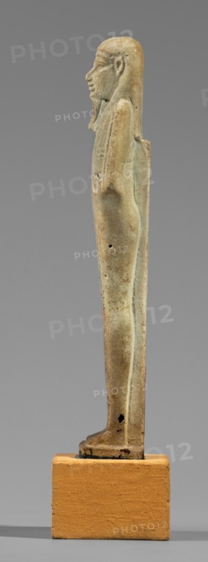 Hornakht's shabti, back pillars, hieroglyphs, wigs, beards facial hair; Hornakht ca. 525–530 BC; funerary figurine, mummy-shaped, standing on a base, back supported by a pillar/pilaster. Arms crossed on the chest, right folded over the left. Right hand holds a long-bladed hoe and two strings from which hangs a semicircular basket on the back, slightly above the elbow; left hand holds a whip. Hands and tools are engraved. Long, three-part smooth wig; long straight beard of uniform width. Face with coarse features, roughly defined eyes, wide nose, prominent mouth with a slight smile. Right ear significantly larger than the left. Inscription carved in a single column on the front from chin to feet, naming the deceased Hornakht and his mother content otherwise unclear. Date: 664–30 BC Late Period–Ptolemaic Period, Egypt; shabti; height 11.6 cm, width 3.1 cm., A slender vertical statuette in muted beige and pale green tones stands on a rectangular ochre base, showing a side profile of a hum...