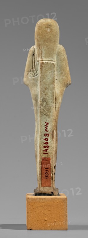 Hornakht's shabti, Back pillars, hieroglyphs, wigs, beards facial hair. Hornakht ca. 525–30 BC. Funerary figurine mummy-shaped shabti, standing on a base, back supported by a pillar/pilaster. Arms crossed on the chest, right folded over the left. In the right hand a long-bladed hoe and two strings supporting a semicircular basket on the back, slightly above the elbow; in the left hand a whip. Hands and tools are engraved. Head wears a long, three-part smooth wig. Beard long, straight, uniform width. Face with coarse features: roughly defined eyes, wide nose, prominent mouth with a slight smile. Right ear significantly larger than the left. Inscription carved in a single column on the front, extending from chin to feet; content unclear, names the deceased Hornakht and his mother. Date: 664–30 BC Late Period–Ptolemaic Period, Egypt. Type: shabti. Height 11.6 cm; width 3.1 cm., A small upright figurine stands on a rectangular base, shown from the back against a neutral gray background. Th...