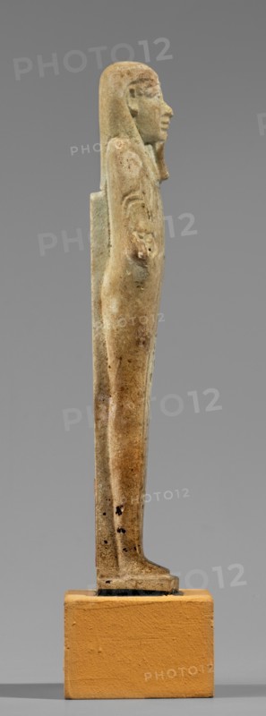 Hornakht's shabti, back pillars, hieroglyphs, wigs, beards facial hair. Hornakht ca. 525–30 BC. Funerary figurine mummy-shaped shabti, standing on a base, back supported by a pillar/pilaster. Arms crossed on the chest, right folded over the left. In the right hand is a long-bladed hoe and two strings, from which hangs a semicircular basket on the back slightly above the elbow; in the left hand a whip. Hands and tools are engraved. Wears a long, three-part smooth wig. Long straight beard of uniform width. Face with coarse features, roughly defined eyes, wide nose, prominent mouth with a slight smile. Right ear significantly larger than the left. Inscription carved in a single column on the front, extending from chin to feet; names the deceased Hornakht and his mother content otherwise unclear. Date: 664–30 BC Late Period–Ptolemaic. Egypt. Type: shabti. Height 11.6 cm, width 3.1 cm., A slender, upright figurine viewed in profile standing on a rectangular plinth, with a smooth elongated f...