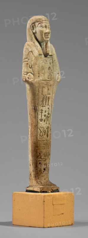 Hornakht's Shabti, back pillars, hieroglyphs, wigs, beards facial hair, Hornakht ca. 525–530 BC, funerary figurine mummy-shaped shabti, grave good, standing on a base, back supported by a pillar/pilaster. Arms crossed on the chest, right folded over the left. In the right hand: a long-bladed hoe and two strings from which hangs a semicircular basket on the back slightly above the elbow; in the left hand: a whip. Hands and tools are engraved. On the head: a long, three-part smooth wig. Long straight beard of even width. Face with coarse features, roughly defined eyes, wide nose, prominent mouth with a slight smile. Right ear significantly larger than the left. Inscription carved in a single column on the front, extending from chin to feet; content unclear but names the deceased Hornakht and his mother. Date: 664–30 BC Late Period–Ptolemaic Period, Egypt. Type: shabti. Dimensions: height 11.6 cm, width 3.1 cm., A small standing figurine with a human face and headdress, shown from a sligh...