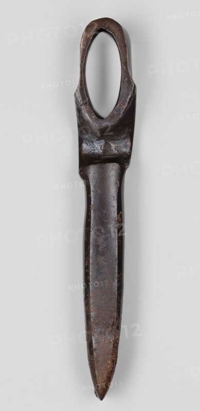 Axe head, unknown Phoenician workshop; Phoenicia Lebanon; 18th–17th century BC. Militaria — edged weapon: axe head with a narrow, elongated blade and a socket for the handle set closer to the butt. Both sides of the socket opening are decorated with a convex shaft in the form of an arch. Weight 610 g., A single elongated metal object photographed against a light gray background, showing a dark brown to nearly black patina with areas of lighter brown rust and wear; the top has an oval opening surrounded by thicker metal, the shaft narrows below that and tapers to a pointed end, with surface irregularities and pitting visible along its length and subtle shine on smoother sections.