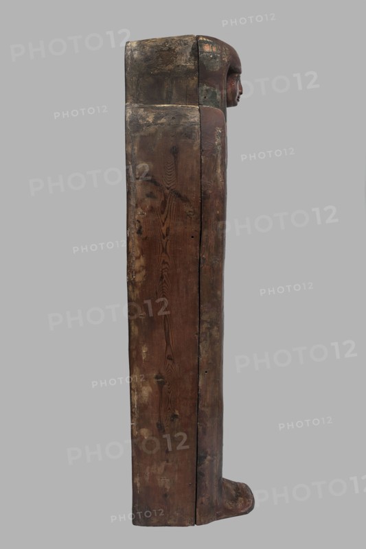 Sarcophagus of the gatekeeper of the temple of Amun Panepi, Panepy ca. 945–715 BC. Sacrificial formulas, necklace iconographic, wig iconographic, mummy straps iconographic, Nut mythological, hieroglyphs, grave goods, sculptured head. A small face with full cheeks painted red. Large eyes with black pupils and white irises. A green wig covering the ears frames the face. A loincloth above the forehead. On the chest is a large useh necklace consisting of nine stripes. On the legs is a vertical strip of the Sacrificial Formula. On the bottom of the chest is an image of Nut marked with a black outline. c. 825–715 BC; Third Intermediate Period, late 22nd Dynasty, Western Thebes. Sarcophagus; height 187 cm, width 49 cm, depth 51 cm., A tall vertical wooden object showing weathered brown and dark brown wood grain with areas of lighter beige wear, a carved human-like profile in reddish-brown at the top right, and traces of darker green and muted ochre paint near the head; the surface has a rough...