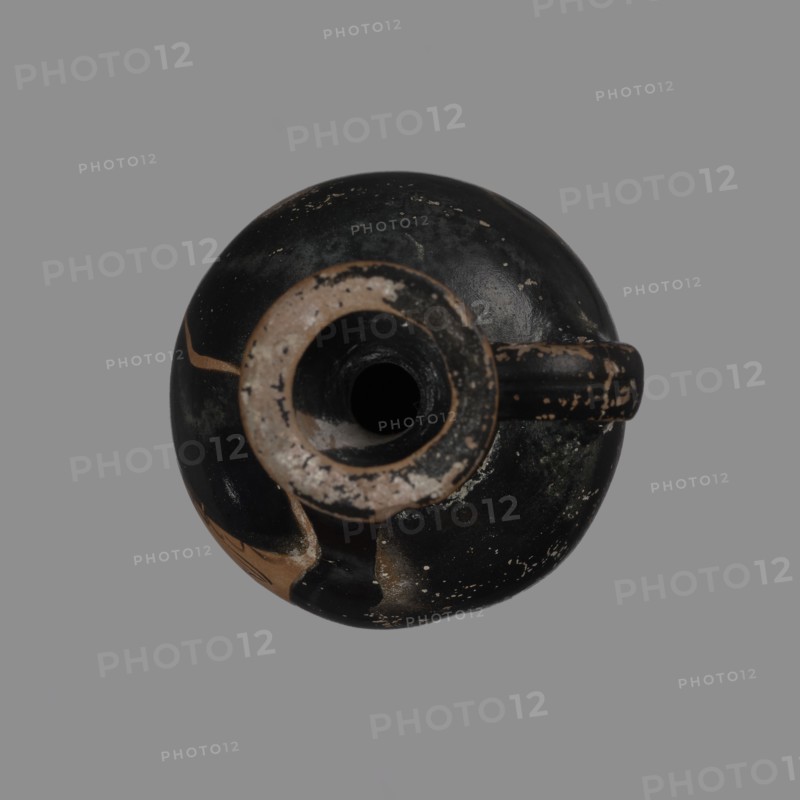 Aryballus lekythos with a ring-shaped foot, a bulging belly, and a high neck widening towards the spout. A rounded vice is attached to the body and the middle of the neck. The vessel is covered with black finish. On the surface of the belly, the head of a woman in a cap turned to the right. Red-figure/black-finished Greek pottery, lekythos aryballus lekythos, late 5th–early 4th century BC. Origin: Attica; finding: Kerch. Dimensions: height 7 cm, width 9 cm, depth 5 cm., A top-down view of a round ceramic vessel against a gray background showing a circular opening and a short handle protruding from the rim; the vessel surface is mostly black with worn areas revealing tan and beige tones, and there are small patches where the black coating is chipped away showing underlying lighter colors; the rim area has a rough beige ring with dark inner shadow, and the handle has similar wear with contrasting black and tan; the overall texture appears weathered with scattered small light flecks.