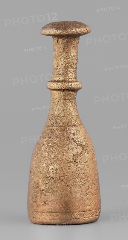 Bottle-type idol base, unknown Luristan workshop. Bottle-shaped base with a long, narrow neck terminating in an upturned, slightly convex spout. Halfway up, a convex cylinder with an engraved zigzag line encircles the neck. Engraved decoration of horizontal bands of line motifs—diagonal crosses, chevrons, hatched triangles—on the central part of the neck and at the lower edge of the base. The base is open at the bottom and hollow inside; a sleeve-shaped hole passes through the neck to allow mounting of a pin with an idol figurine. Geometric ornaments; bottle base. Iron Age II, 8th–7th century BC. Luristan, Iran. Base; height 7.5 cm, width 2.8 cm., A single sculpted object resembling a bottle or small vessel stands upright against a plain grey background; the object has a wide rounded base that tapers into a narrower neck topped by a flattened, mushroom-like rim and a pronounced collar around the midneck. The surface appears rough and textured with visible pitting and irregularities, an...