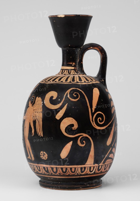 Red-figure aryballus lekythos with a scene of Aphrodite's toilet, On the belly: Aphrodite crouching and Eros handing her an alabastron, beginning of the 4th century BC previously dated to the 2nd quarter of the 4th century BC, findspot Nola, origin Campania, vessel: lekythos aryballus lekythos, height 16.5 cm, width 19.5 cm, weight 0.23 kg, subjects: Aphrodite, Eros, Greek deities., A black ceramic vessel with a rounded body, narrow neck, flared rim, and a single handle, decorated with orange-colored painted figures and swirling motifs; the upper neck area shows a band of vertical petal-like shapes in orange, the body features large scrolling tendrils and teardrop forms in orange against the black background, and a partial human-like figure with wings rendered in orange is visible on one side; the base has a repeating orange decorative border and the rim and handle show small areas where the black surface is chipped, revealing an orange underlayer.