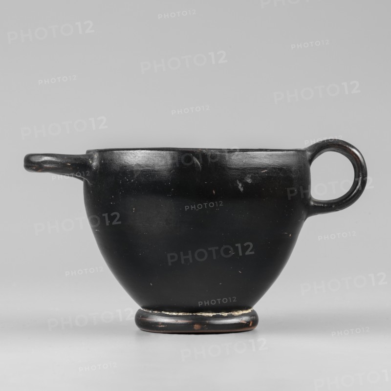 Black-finished skyphos, Greek ceramics, Type B. Thin-walled skyphos with a deep bowl and annular foot. One handle mounted horizontally, the other vertically. Entirely covered with a high-quality, glossy black varnish; only the bottom remains clay-colored with decoration of concentric circles. Technology: visible traces of turning; small chips on the surface. Clay color 2.5 YR 5/6 red. Varnish color 10 YR 2/1 black. Attributed to Attica, 480–450 BC. Vessel skyphos. Height 7.6 cm; width 14.1 cm; depth 8.8 cm; weight 0.102 kg., A black ceramic cup with a rounded body and a small pedestal base sits against a neutral gray background, showing a simple loop handle on one side and a short spout-like extension on the opposite side; the cup's surface is mostly smooth with a few small scuffs and lighter spots, and there are subtle variations of black and dark brown on the rim and base.