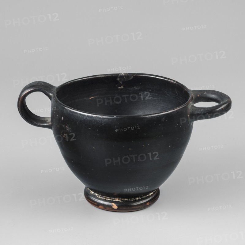 Black-finished skyphos, Type B. Thin-walled skyphos with a deep bowl and annular foot. One handle mounted horizontally, the other vertically. The object is entirely covered with a high-quality, glossy black varnish; only the bottom remains clay-colored, decorated with concentric circles. Technology: visible traces of turning; small chips on the surface. Clay color: 2.5 YR 5/6 red. Varnish color: 10 YR 2/1 black. Attributed to Attica, 480–450 BC. Vessel skyphos. Height 7.6 cm; width 14.1 cm; depth 8.8 cm; weight 0.102 kg., A black ceramic cup with a rounded body and a low foot, featuring a pair of horizontal loop handles on opposite sides, sits against a light gray background; the cup shows small worn spots and areas where the black finish is chipped to reveal a brownish color underneath, and the interior and rim display subtle scuffs and lighter marks.