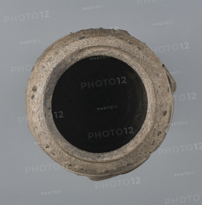 Vise-free cup, unknown Rhine workshop, Goluchów Greater Poland Voivodeship. Hard-fired, granular, light gray clay 7.5YR N7/; gray matte glaze 10YR 5/18 covering entire surface, intentionally rough sandblasted. Upper body with en barbotine decoration of stylized lanceolate leaves between two stripes of dots. Whole vessel, 2nd half of the 1st century, Cologne, height 8.0 cm, width 7.0 cm., A top-down view of a round clay vessel with a wide dark interior and a rough textured rim showing irregularities and small protrusions, in shades of beige, tan, gray, black, and brown against a muted gray background.