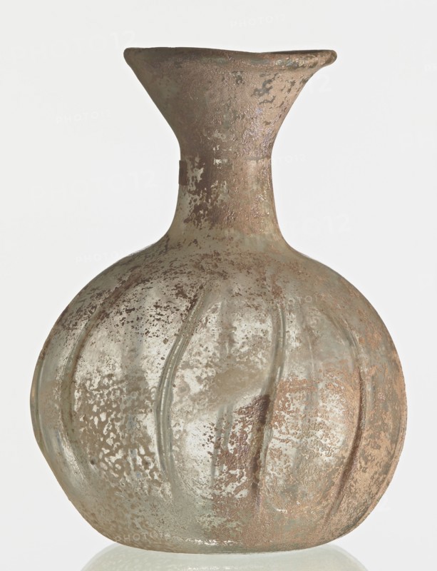 Flask; body in the shape of a flattened sphere, decorated with ribs;, Syria; 2nd–3rd century AD. Transparent, colourless, iridescent glass. Slightly flattened spherical body decorated with vertical thin, uneven ribs; short cylindrical neck terminating in a funnel-shaped spout with a thickened and rounded edge; free-blown. Outer diameter of the spout: 3.80 cm. Height: 8.80 cm. Body diameter: 8 cm. Bottom diameter: 3.40 cm. Type: Isings form 101? Vessel; weight 0.030 kg., A round, bulbous glass vessel with a flared neck and narrow shoulder, showing a textured, mottled surface; the glass appears cloudy and weathered with patches of rough encrustation and smooth areas, and features shallow vertical ridges along the body; visible colors include pale green, muted brown, gray, and hints of beige.