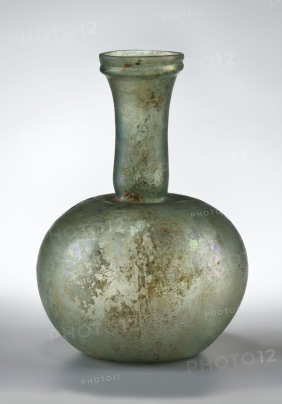 Flask with a spherical body and a high, cylindrical neck ending in a wide, straight-cut rim. The neck is marked by a recess and is decorated with a ring under the rim. Concave bottom with rounded edges. The neck is set obliquely to the body. Translucent, green glass. Spherical body, cylindrical neck slightly flaring upwards, straight rim with rounded edge, heat-worked. Neck narrowed at the bottom; free blowing. Type: Isings form 104B, 3rd–4th century, Phoenicia; vessel. Dimensions: height 18.4 cm; width 13.2 cm; rim diameter 4.9 cm; foot diameter 6 cm; maximum diameter 11.9 cm., A glass vessel with a rounded body and a long narrow neck topped by a slightly flared rim, showing an aged, weathered surface with iridescent sheens and areas of mottled patina; the visible colors include pale green, muted blue, hints of brownish rust, and subtle purple and silver reflections, all set against a neutral gray-white background.