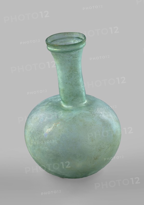 Flask with a spherical body and a high, cylindrical neck ending in a wide, straight-cut rim. The neck is marked by a recess and is decorated with a ring under the rim. Concave bottom with rounded edges. The neck is set obliquely to the body. Translucent, green glass. Spherical body, cylindrical neck slightly flaring upwards, straight rim with rounded edge, heat-worked. Neck narrowed at the bottom; free blowing. Type: Isings form 104B, 3rd–4th century, Phoenicia, vessel. Height 18.4 cm; width 13.2 cm; rim diameter 4.9 cm; foot diameter 6 cm; maximum diameter 11.9 cm. Maker: unknown., A rounded glass vessel with a bulbous body and a long narrow neck topped by a flared rim, shown against a plain neutral background; the glass surface is predominantly a pale green with variations toward bluish-green and hints of turquoise, and it shows iridescent patches and subtle cloudy areas, along with small spots and mottled discoloration in darker green and brown tones, and the interior rim and neck d...
