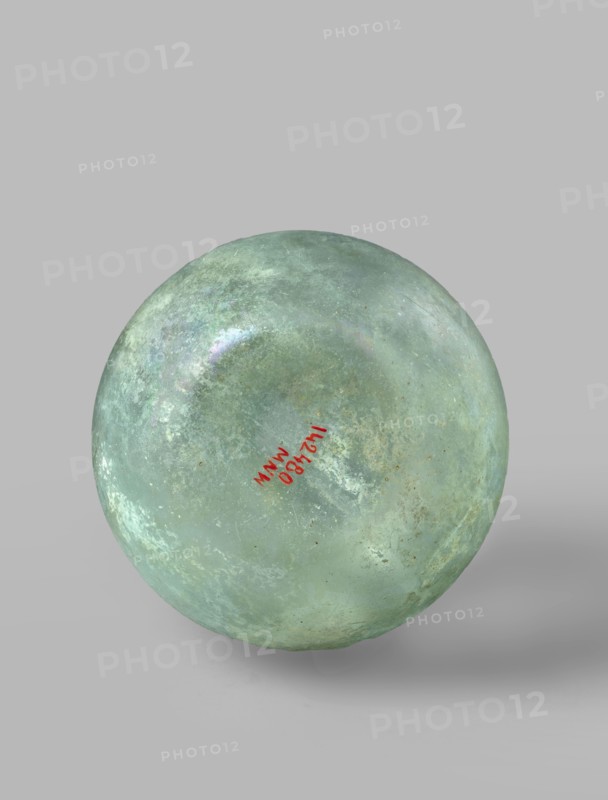 Flask with a spherical body and a high, cylindrical neck ending in a wide, straight-cut rim. The neck is marked by a recess and is decorated with a ring under the rim. Concave bottom with rounded edges. The neck is set obliquely to the body. Translucent, green glass. Spherical body, cylindrical neck slightly flaring upwards, straight rim with rounded edge, heat-worked. Neck narrowed at the bottom; free blowing. Type: Isings form 104B, 3rd–4th century, Phoenicia; vessel. Dimensions: height 18.4 cm; width 13.2 cm; rim diameter 4.9 cm; foot diameter 6 cm; maximum diameter 11.9 cm., A spherical object with a weathered, mottled surface in shades of green and aqua with areas of pale turquoise and faint mottlings of whitish and grey; the sphere has a slightly iridescent sheen in places and small spots of darker green and brownish discoloration; it sits against a smooth light grey background and casts a soft shadow beneath it.