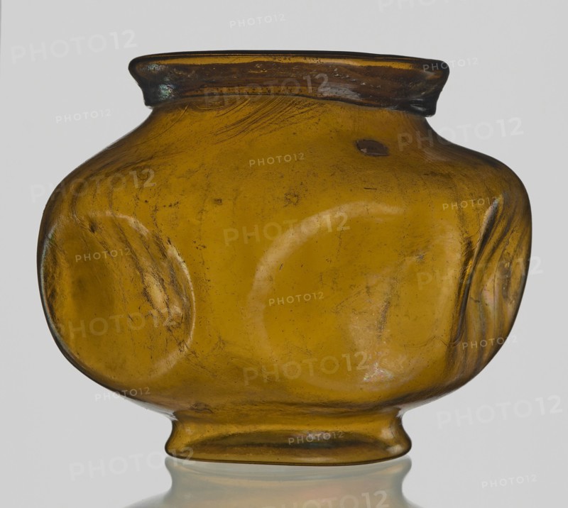 Vessel, Goluchów Greater Poland Voivodeship, vessel, height 10.2 cm, width 13.2 cm, An amber-colored glass vessel with a rounded, bulbous body tapering to a small foot and a slightly flared neck, showing a few shallow circular indentations on its sides, visible surface scratches and small bubbles within the glass, and areas of darker brown near the rim and subtle iridescent sheens where the light catches the surface; the background is neutral light gray.