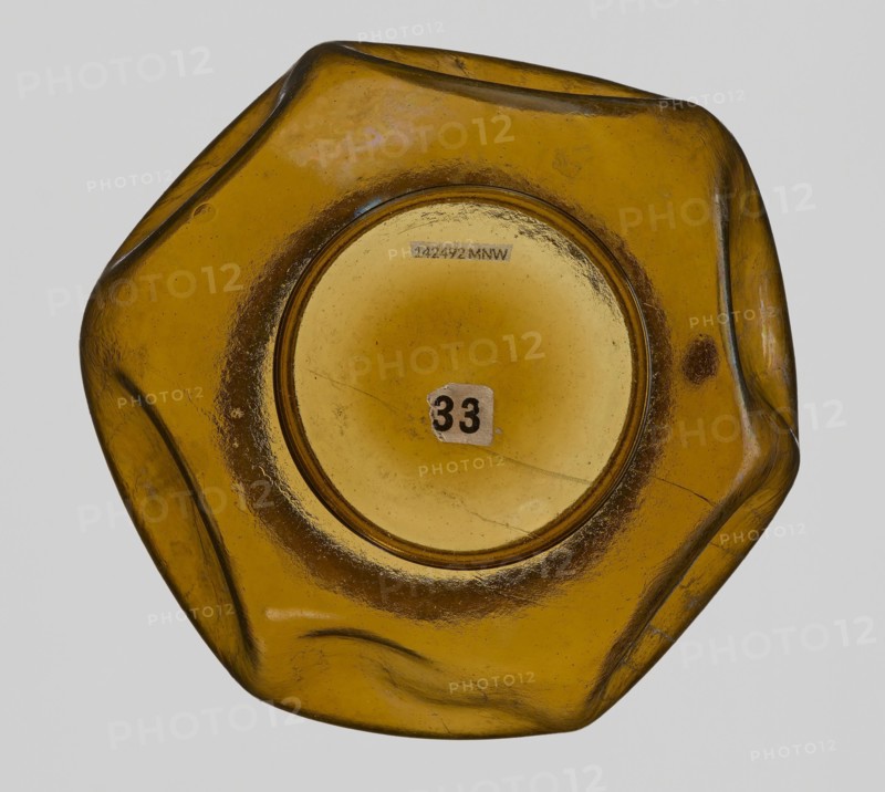 Vessel, Goluchów Greater Poland Voivodeship, vessel, height 10.2 cm, width 13.2 cm, A close-up view of a translucent amber-colored glass object with an irregular hexagonal shape and smooth, rounded edges, showing a central circular recessed area and a small worn sticker; the surface has variations in color from golden yellow to darker brown tones and shows scratches, scuffs, and a few small bubbles or imperfections.