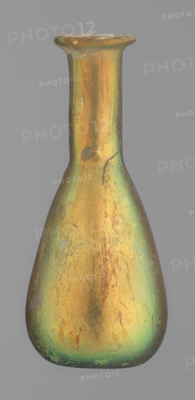 Flask, unknown eastern workshop, glass, antiquity, Roman Empire. Flask with a slender, pear-shaped body and a lower neck ending in a ring-shaped rim. Flat bottom. Covered with multicolored patina. 1st–2nd century, vessel, height 9.4 cm, A single narrow-necked glass vessel with a flared rim and a bulbous body sits against a plain gray background; the surface shows an iridescent finish with predominant gold and green hues, along with areas of muted bronze and hints of brown, uneven texture, small mottled spots, and patches of wear that reveal subtle variations in sheen and color across the body.