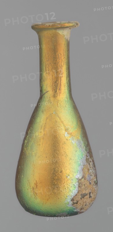 Flask, unknown eastern workshop, Goluchów Greater Poland Voivodeship, glass, antiquity, Roman Empire. Flask with a slender, pear-shaped body and a lower neck ending in a ring-shaped rim. Flat bottom. Covered with multicolored patina. 1st–2nd century, vessel, height 9.4 cm, A narrow-necked bottle with a flared rim, shown against a plain gray background, displaying an iridescent surface with dominant gold and green tones and areas of worn or rough texture in muted gray and brown.